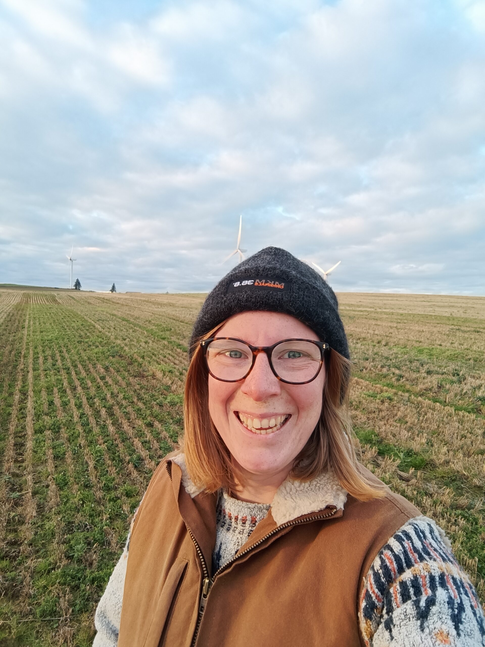 A smiling person wearing glasses, a knit hat, and a brown vest stands in a grassy field with wind turbines and a cloudy sky in the background.