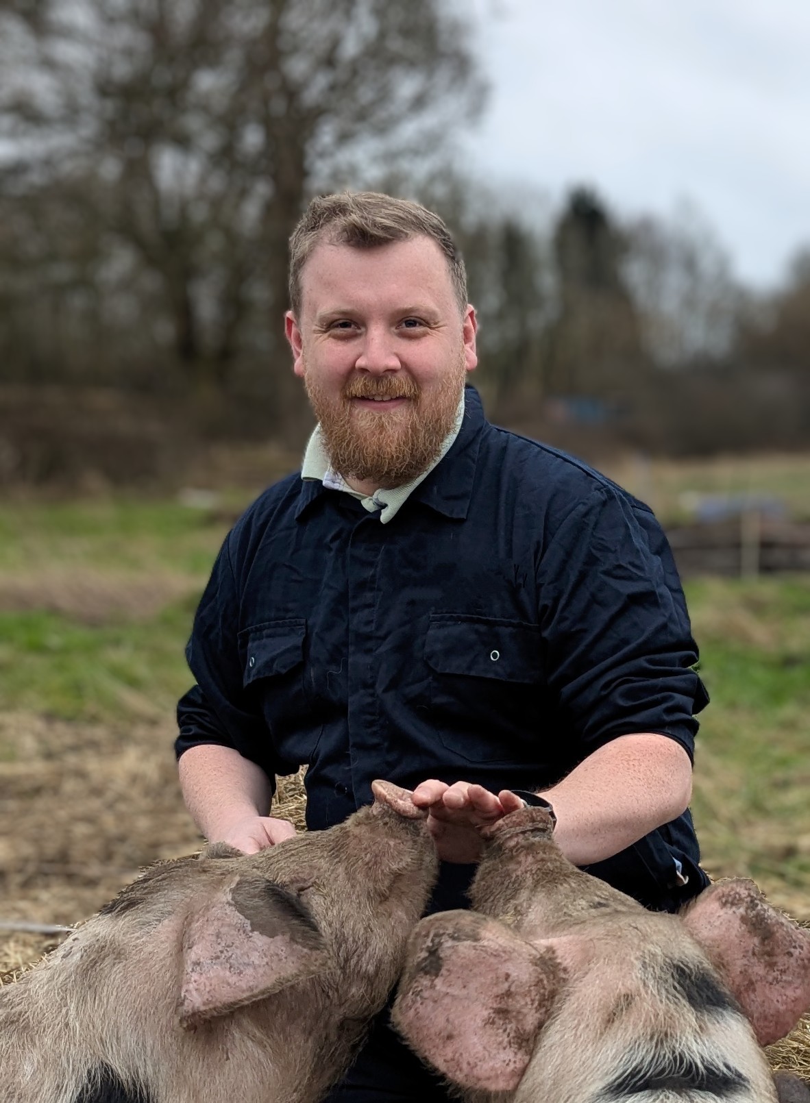 A man with a beard in a navy jacket smiles while kneeling outdoors, gently petting two pigs. The background features grassy fields and leafless trees under a cloudy sky.