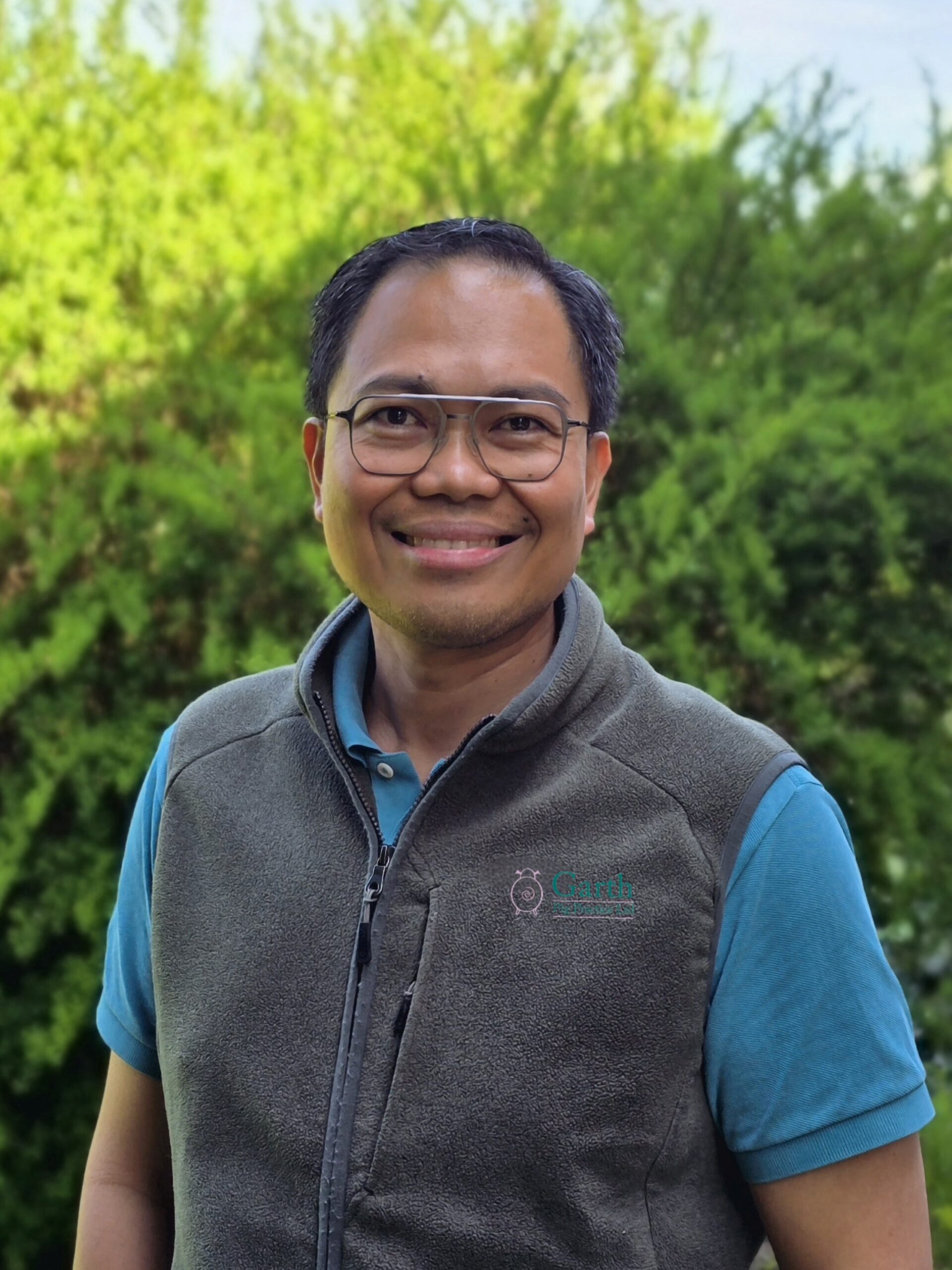 A smiling person wearing glasses, a blue shirt, and a gray vest stands outdoors in front of lush green bushes on a sunny day.